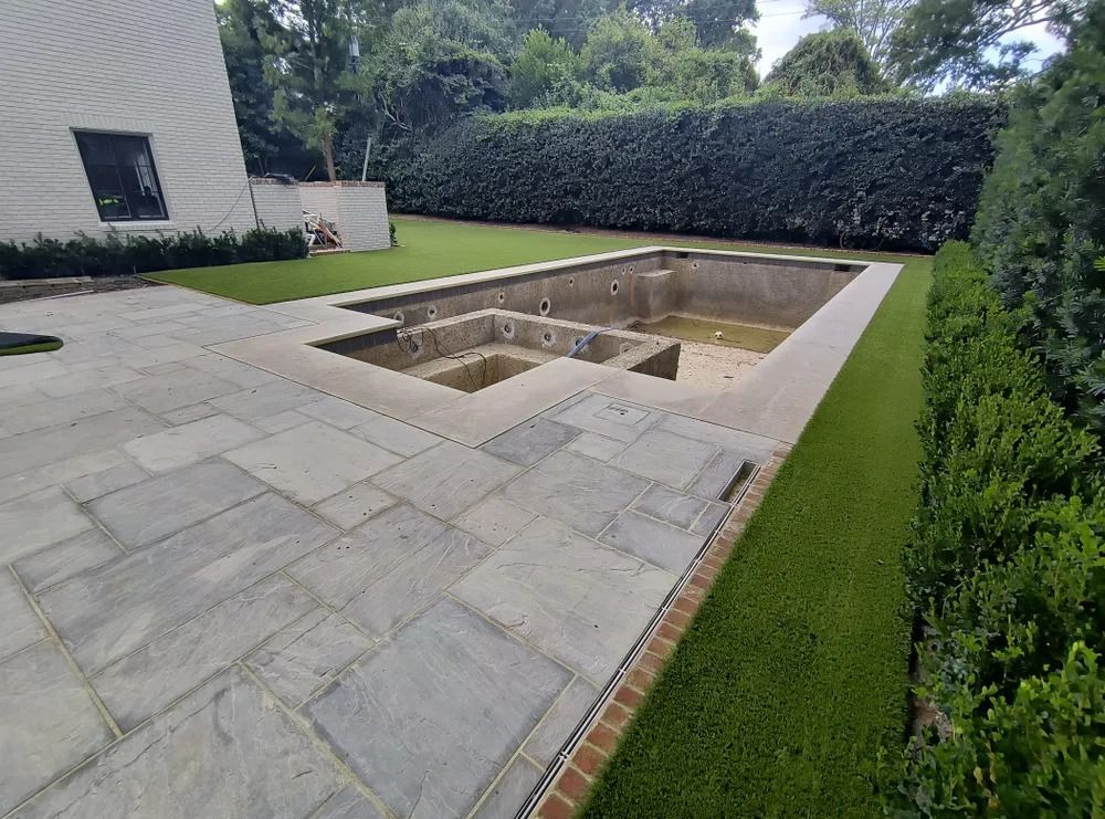 Empty rectangular backyard pool under construction beside a patio and hedge-lined lawn