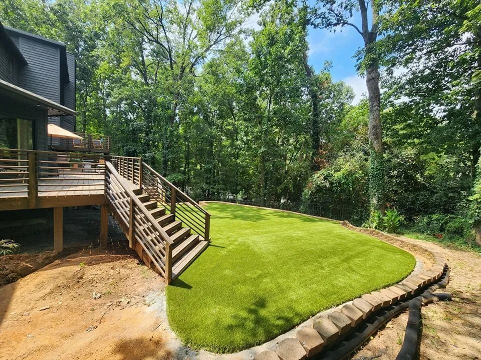 Backyard with wooden deck stairs, bright green lawn, and wooded landscaping beside a house