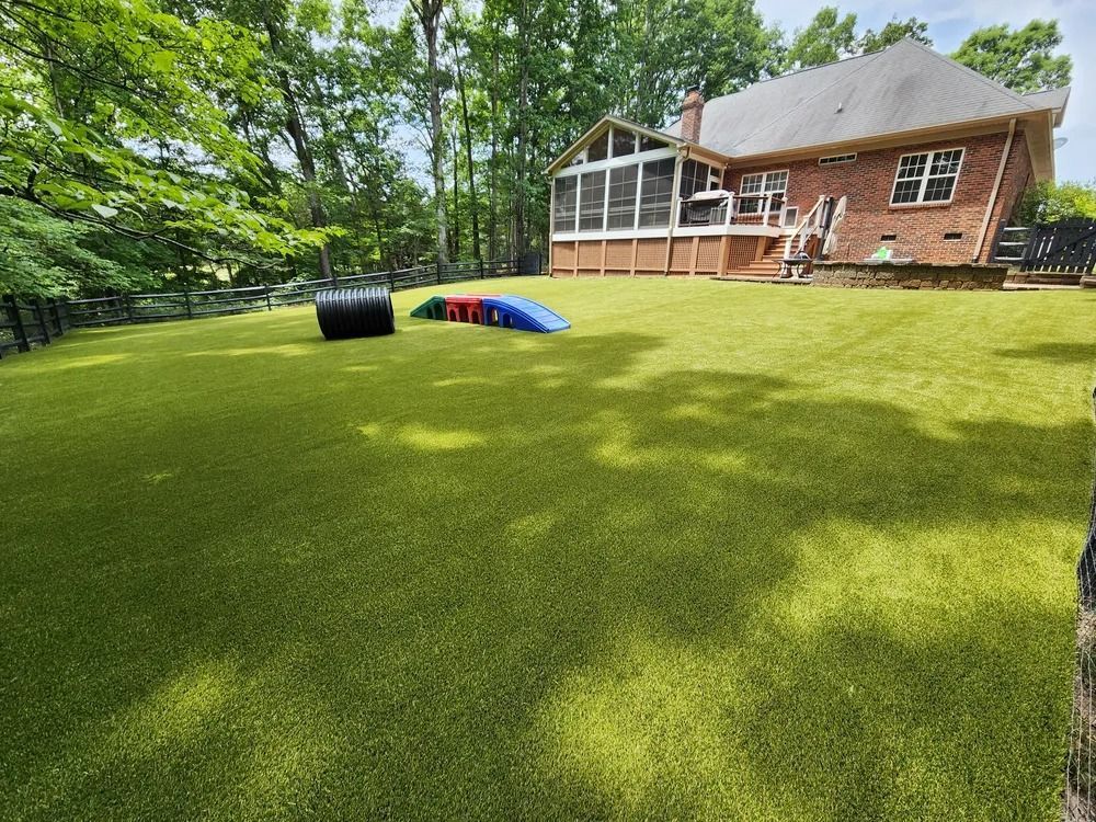 Backyard with bright green lawn, trees, and a brick house; colorful play tunnel near the patio.