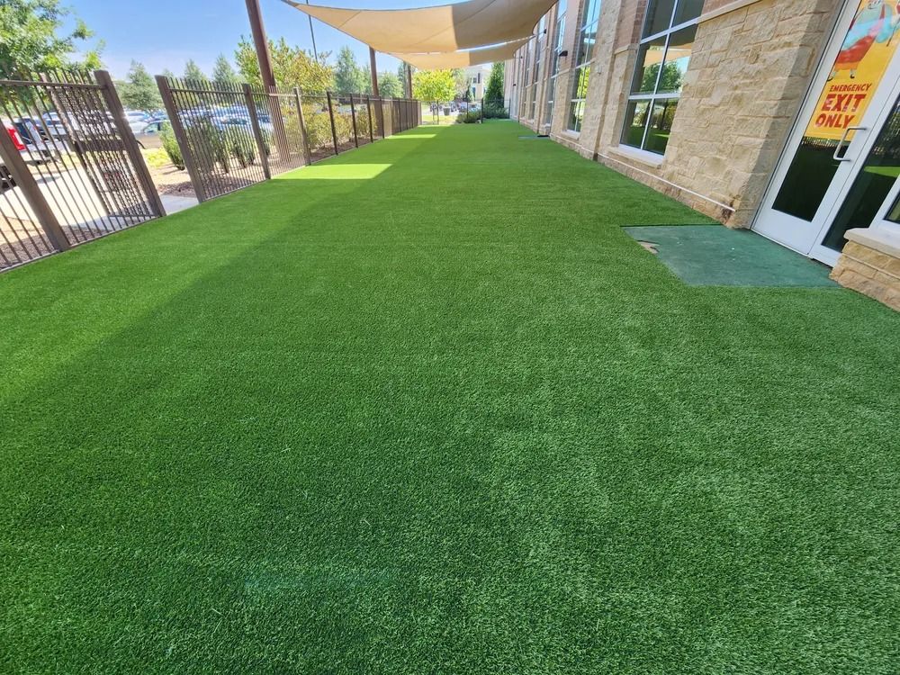 Covered walkway with artificial green turf beside a building and fenced sidewalk.