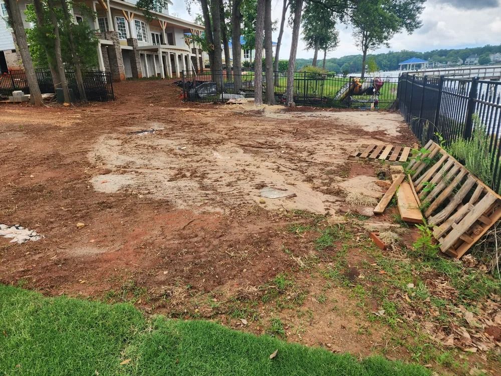 Bare dirt lot by a lakeside fence, with trees, a building, and stacked wooden pallets.