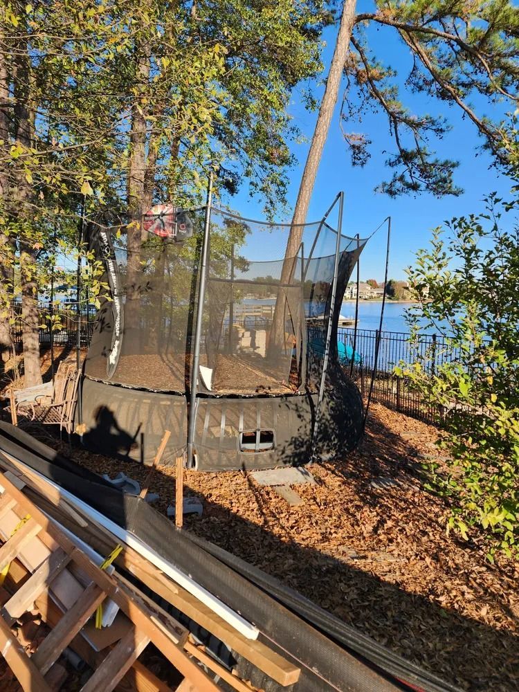Backyard trampoline with safety net beside a house, surrounded by trees and fallen leaves.