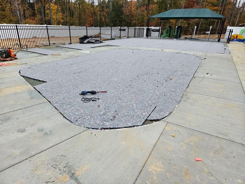 Empty concrete pool with a large gray patch, outdoors beside a fence and pavilion.