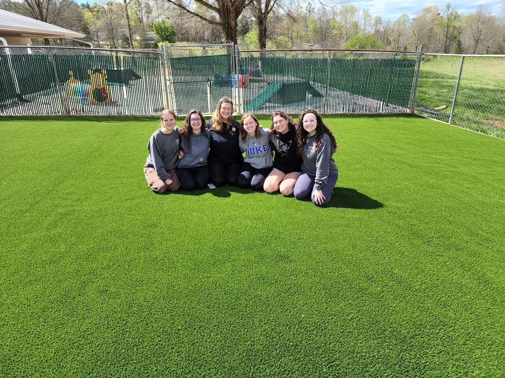 Five people sitting on bright green turf in front of a mural and fence, smiling outdoors.