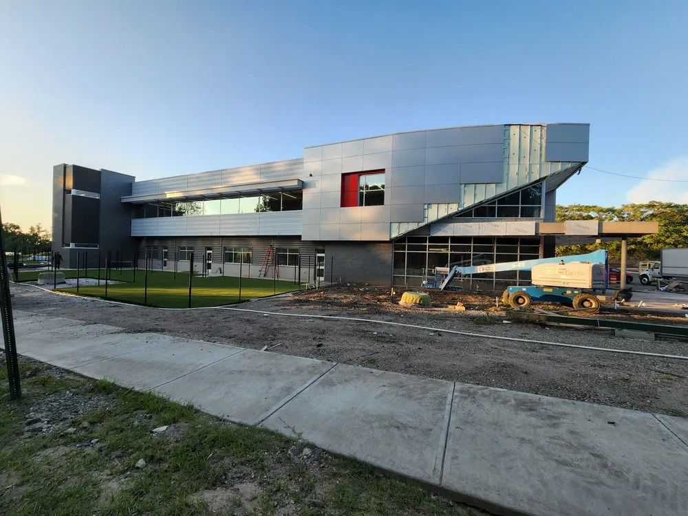 Modern building under construction with metal facade, fenced site, and dirt work area at sunset