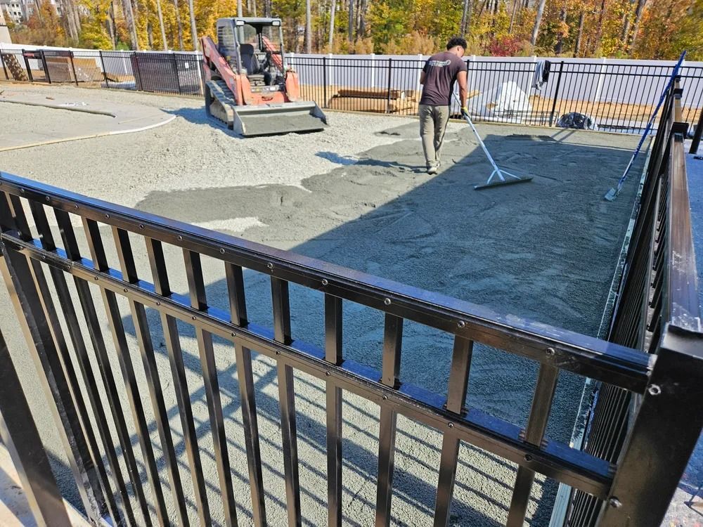 Workers leveling wet concrete in a fenced outdoor construction area with a skid steer nearby