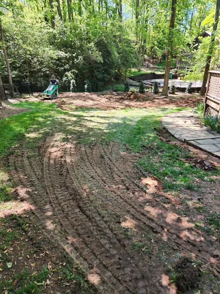 Freshly tilled backyard soil with curved tracks, bordered by grass and trees, beside a patio and deck