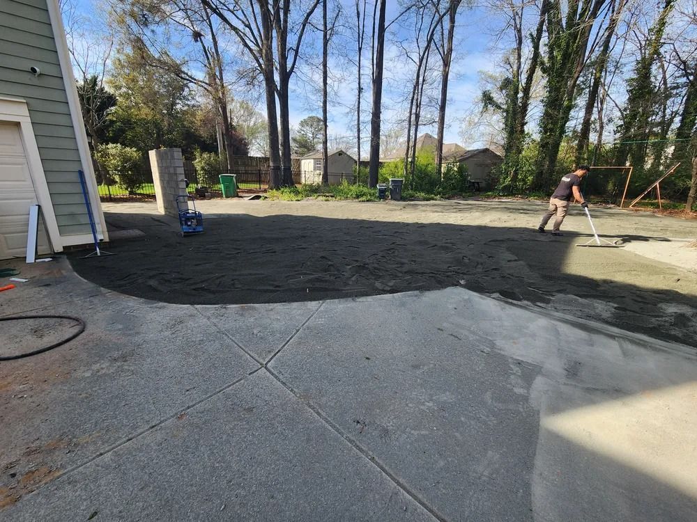 Backyard with freshly laid dark soil beside a concrete patio; a person works near the right edge.