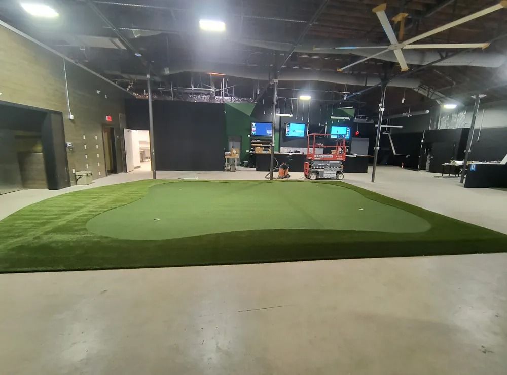 Indoor golf practice area with green turf mat, nets, and screens in a dimly lit facility