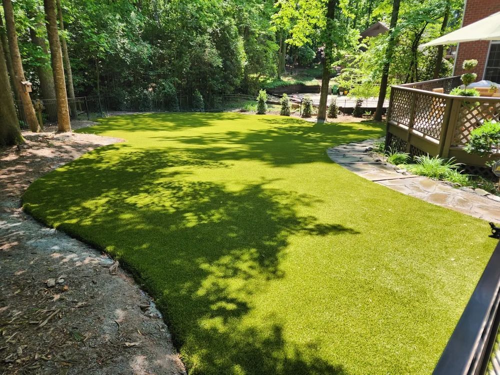 Curved green lawn in a shaded backyard with trees, a stone path, and a wooden deck railing.