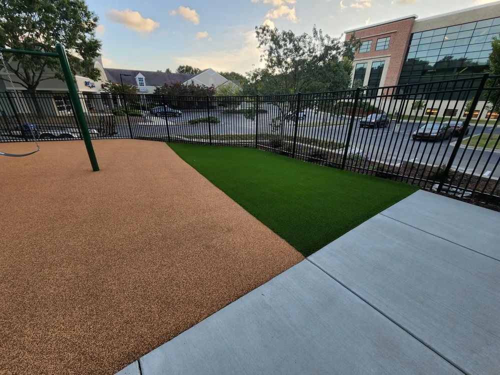 Small fenced outdoor area with tan rubber surface, green turf, and concrete walkway beside buildings.