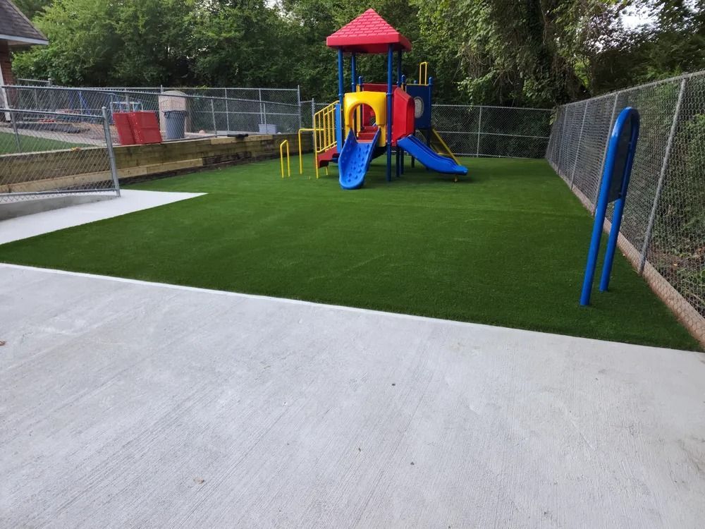 Empty playground with colorful playset on green turf beside a paved area and chain-link fence