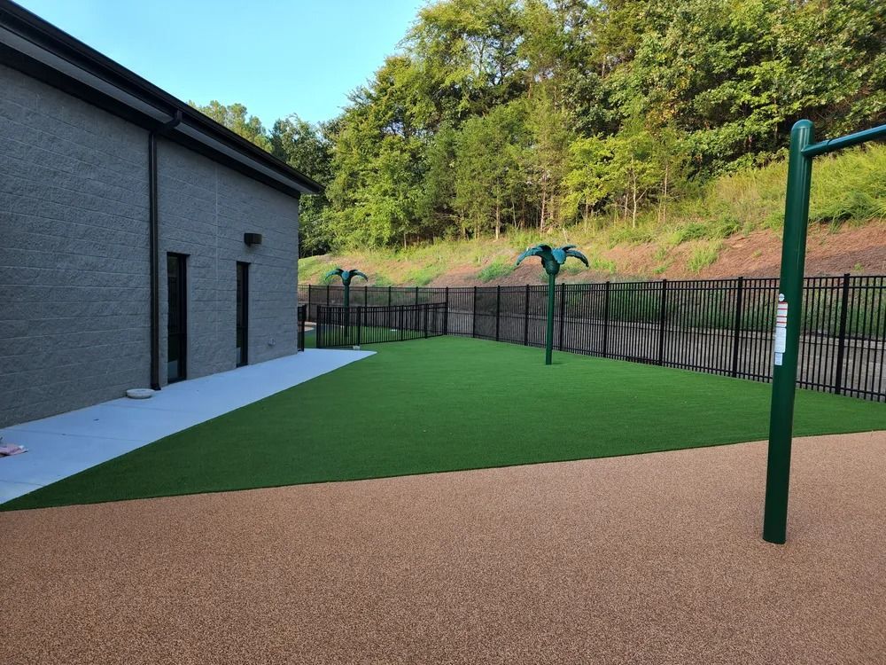 Backyard with artificial turf, tan ground cover, a gray building, and a fence along a wooded slope