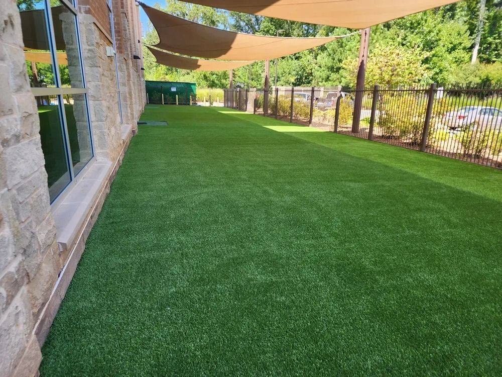 Covered artificial turf patio beside a brick building, shaded by tan sails and bordered by a fence and greenery