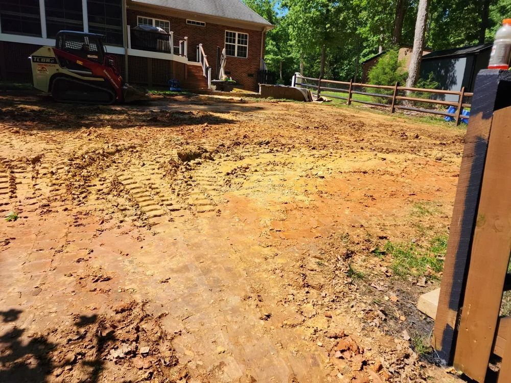 Muddy, recently graded yard beside a brick house, with a small tractor and fence in the background
