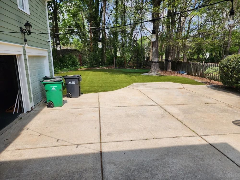 Concrete driveway beside a green garage, with trash bins and a grassy backyard bordered by trees.