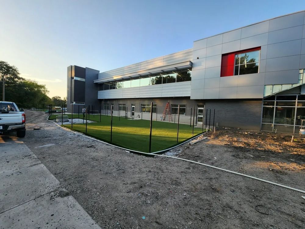 Modern building exterior with a fenced green courtyard and gravel construction area at sunset