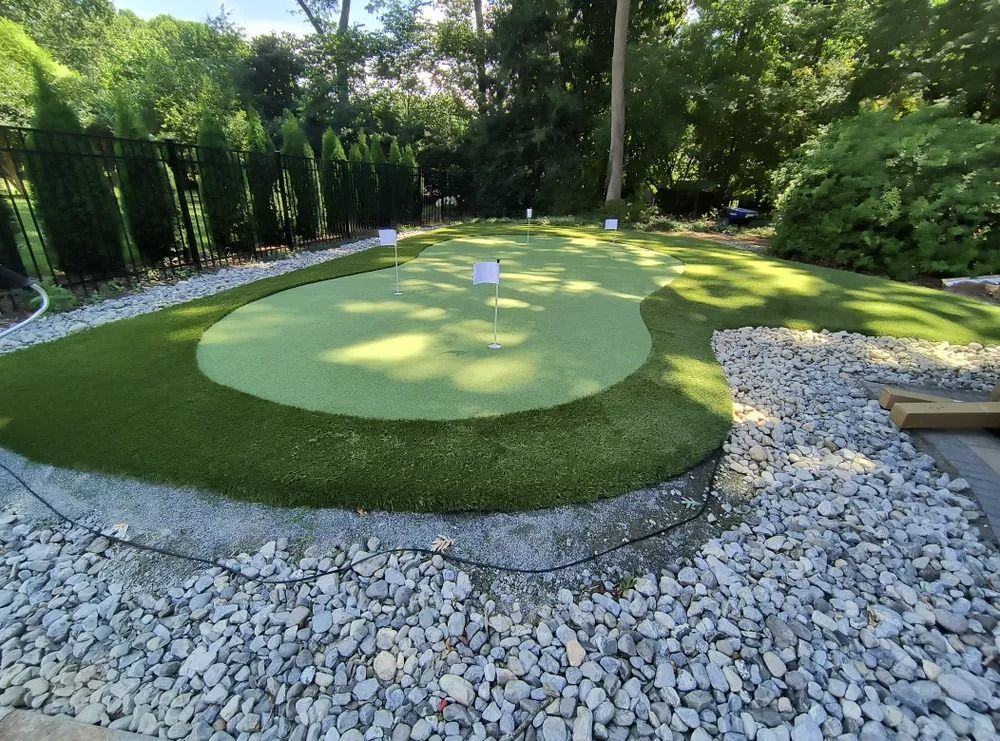 Backyard putting green with a golf hole, bordered by white stones and surrounded by trees