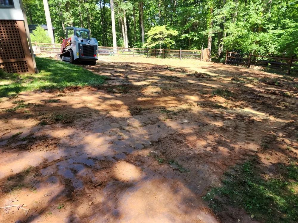 Muddy yard with a blue tractor near a wooden shed, surrounded by trees and fence shadows