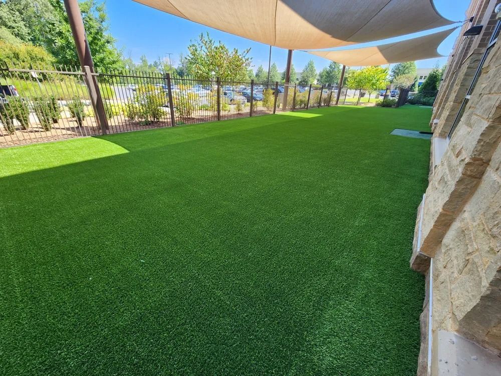 Shaded artificial turf lawn beside a stone wall and garden with trees and tan shade sails