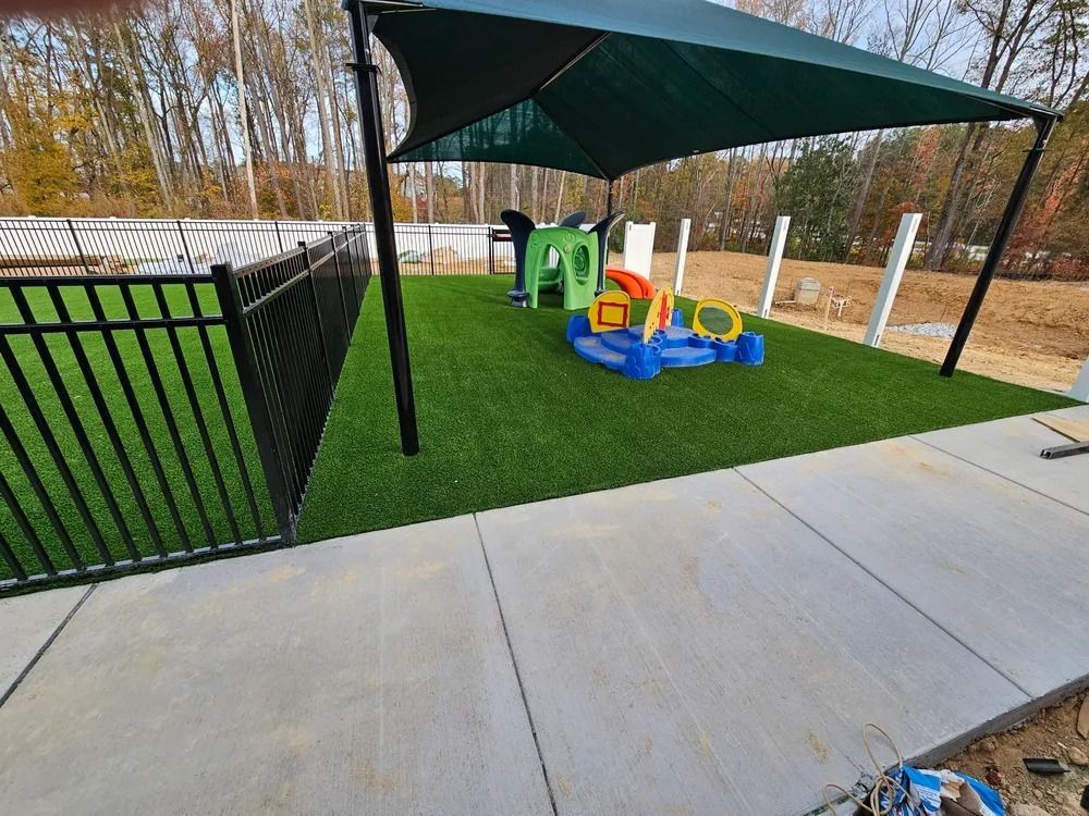 Covered playground with small play car on green turf beside a concrete path and black fence.