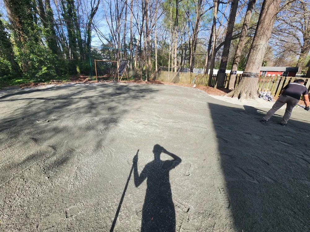 Gravel lot with trees and a person walking at right; long shadow of the photographer in the foreground