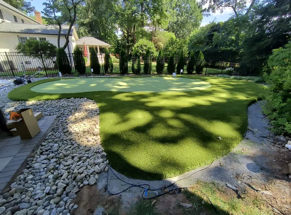 Backyard with freshly laid green lawn, stone border, and lush trees beside a house and patio umbrella