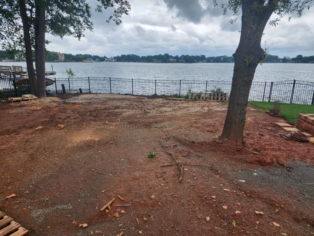 Dirt clearing by a lakeshore with trees, a fence, and calm water under an overcast sky