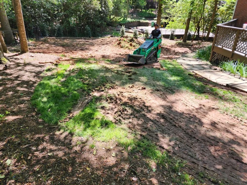 Backyard with a green lawn tractor on a patchy dirt and grass slope near trees and a deck