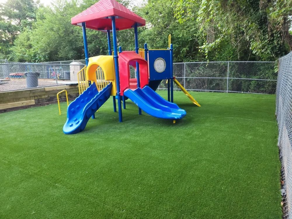 Colorful playground with blue slides, red roof, and green artificial turf beside a chain-link fence.