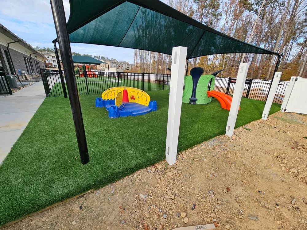 Outdoor playground with artificial turf, shade sail, and colorful play equipment beside a fenced area