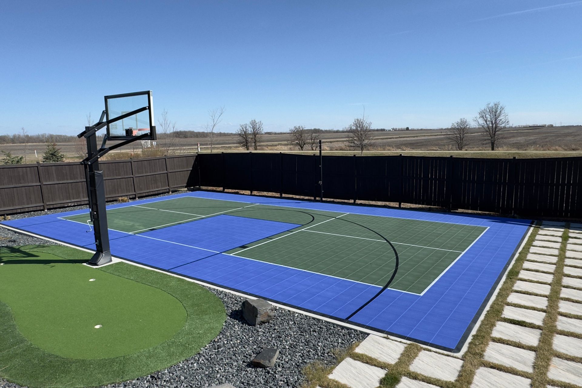 Outdoor sports court with blue and green surfaces, a hoop, and a black fence under a clear sky