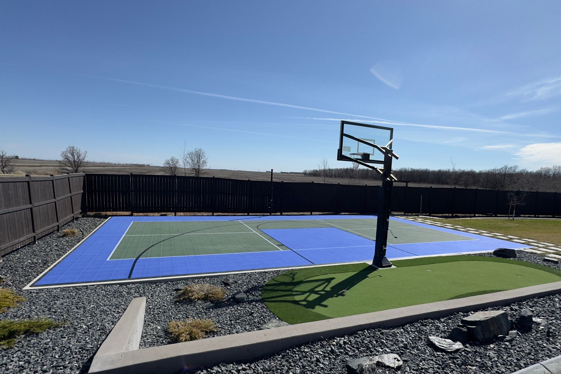 Outdoor basketball court with blue and green surfaces and a single hoop, surrounded by fencing under a clear sky