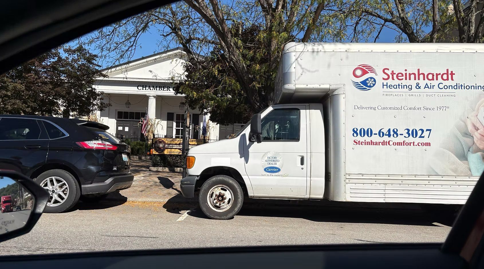 Steinhardt delivery truck parked outside a home beside a black SUV, with a patio and trees in front.