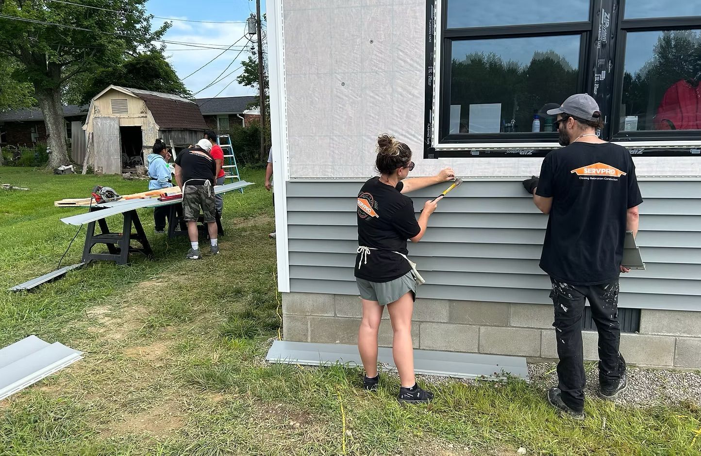 People measuring gray siding on a house while others work at a table in the yard.