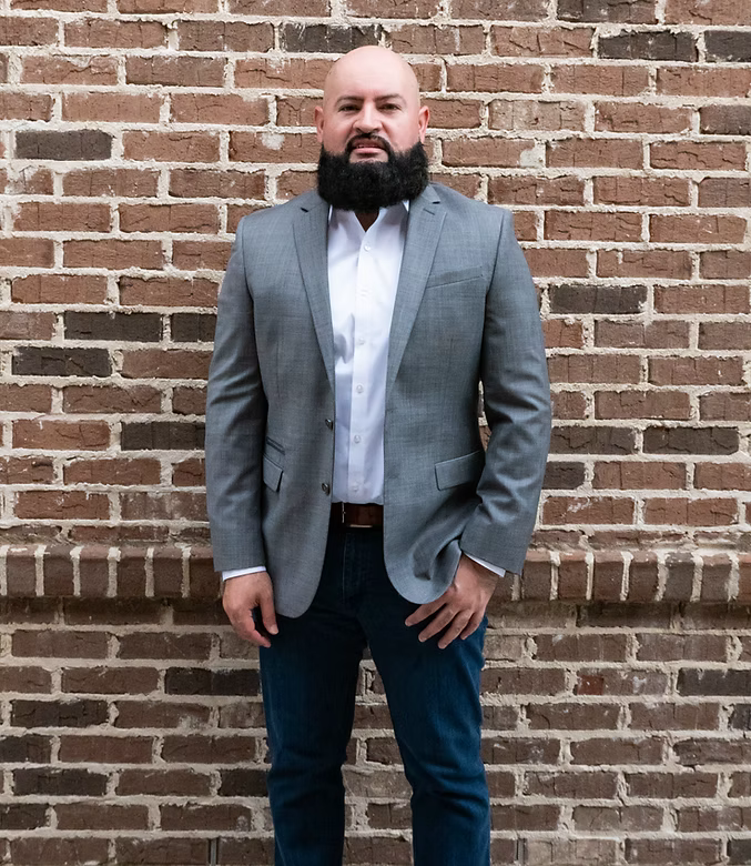 Man standing against a brick wall, wearing a gray blazer, white shirt, and dark jeans.