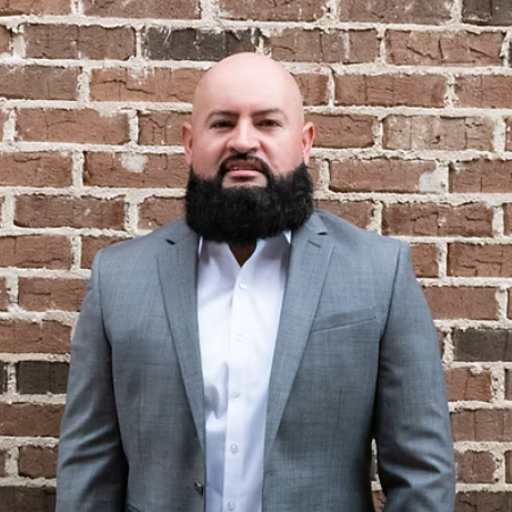 Man in a gray suit and white shirt standing against a brick wall, facing the camera.