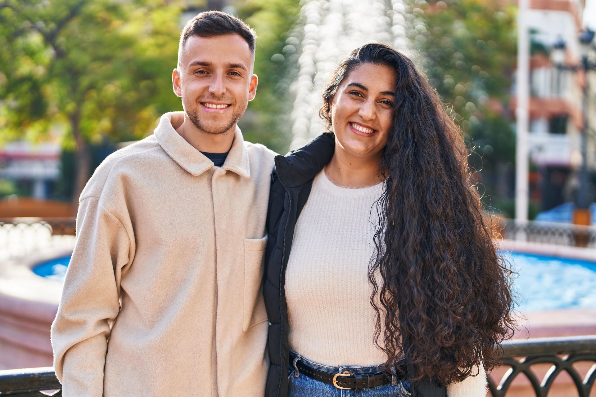 Two smiling people standing outdoors near a fountain on a sunny day