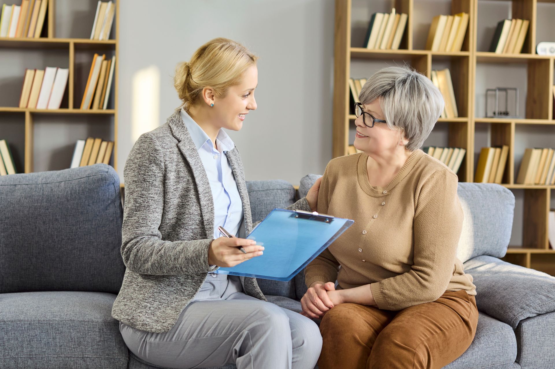 Two women sit on a couch, talking with a clipboard in a bright living room.