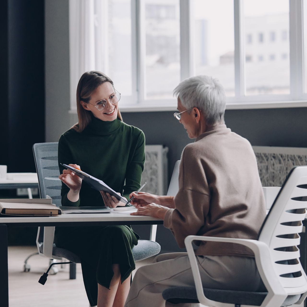 Two people talk at a table in a modern office, one holding a clipboard and pen.
