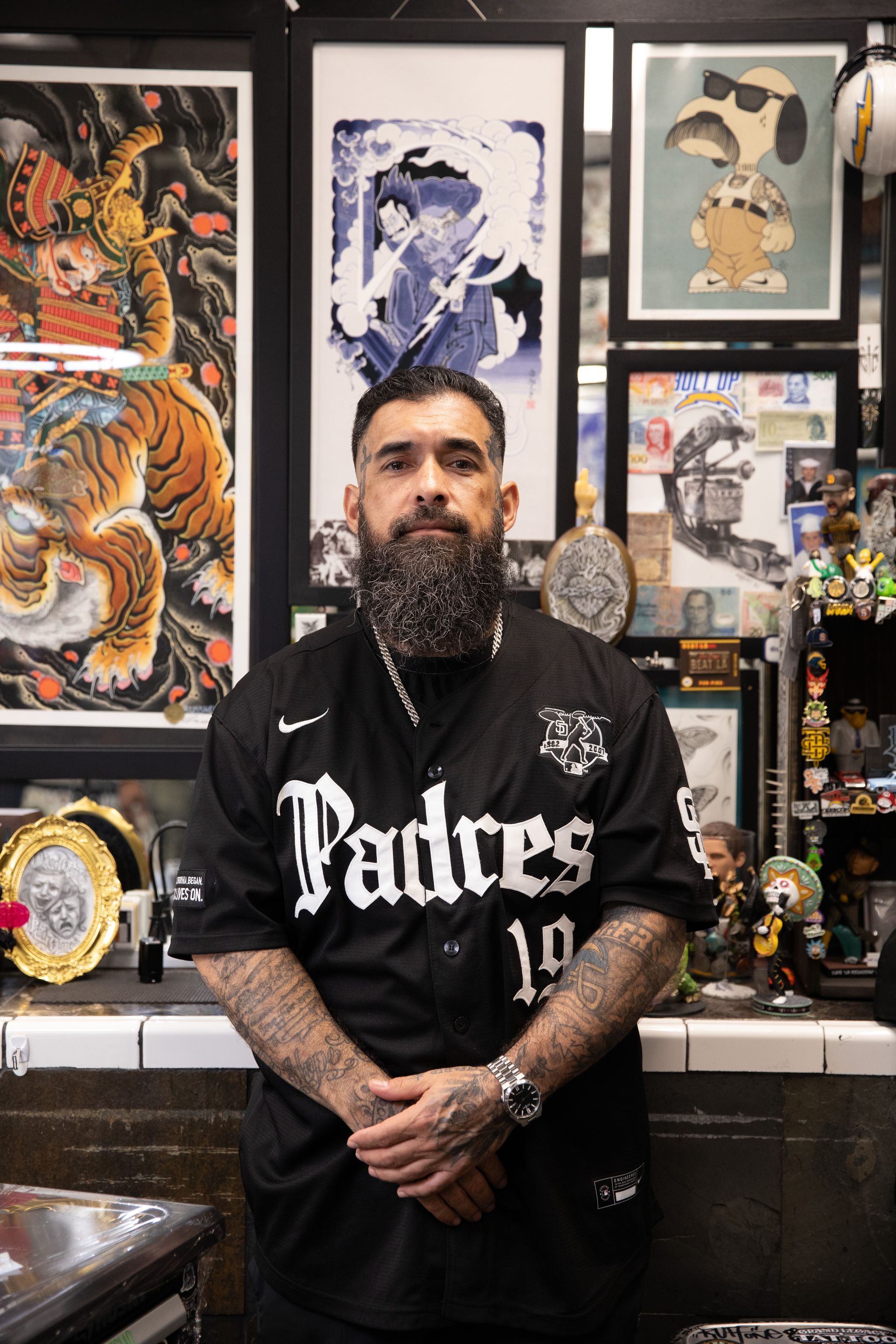 Man standing in a Padres jersey in front of colorful framed art and collectibles, arms crossed.