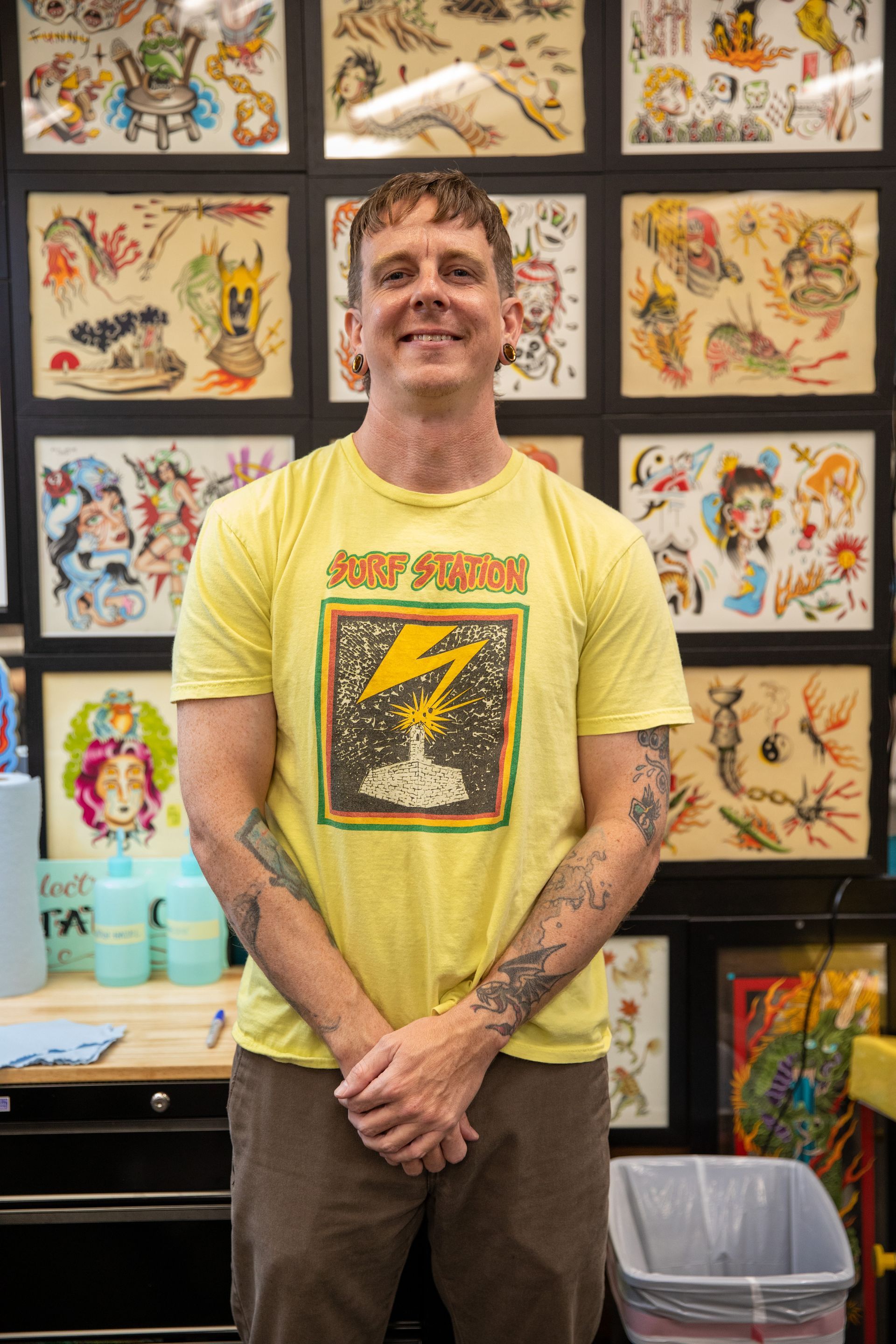 Smiling person in a yellow graphic T-shirt standing in front of a wall of framed artwork and a counter