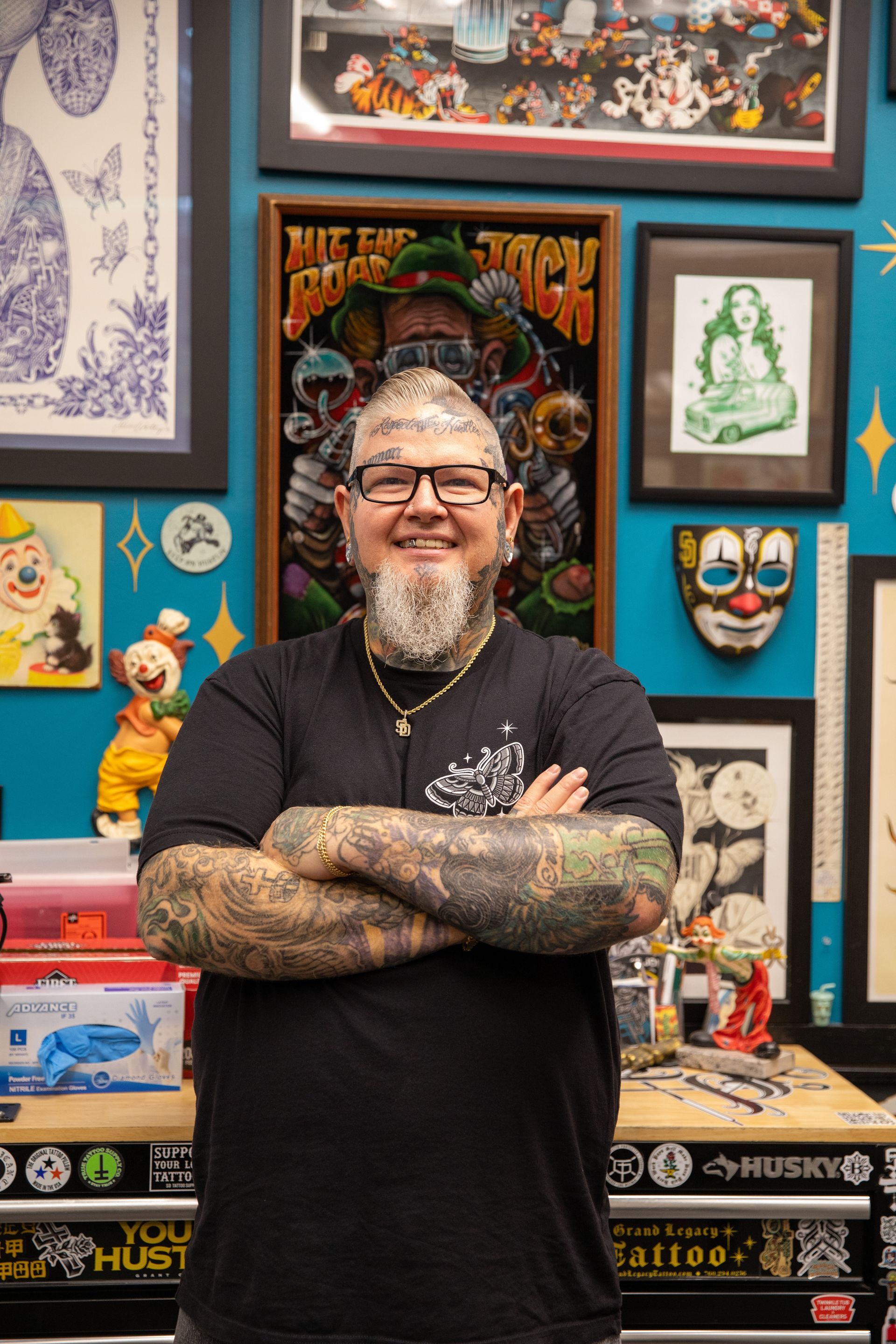 Smiling bearded man with tattooed arms stands in a colorful art studio, arms crossed.