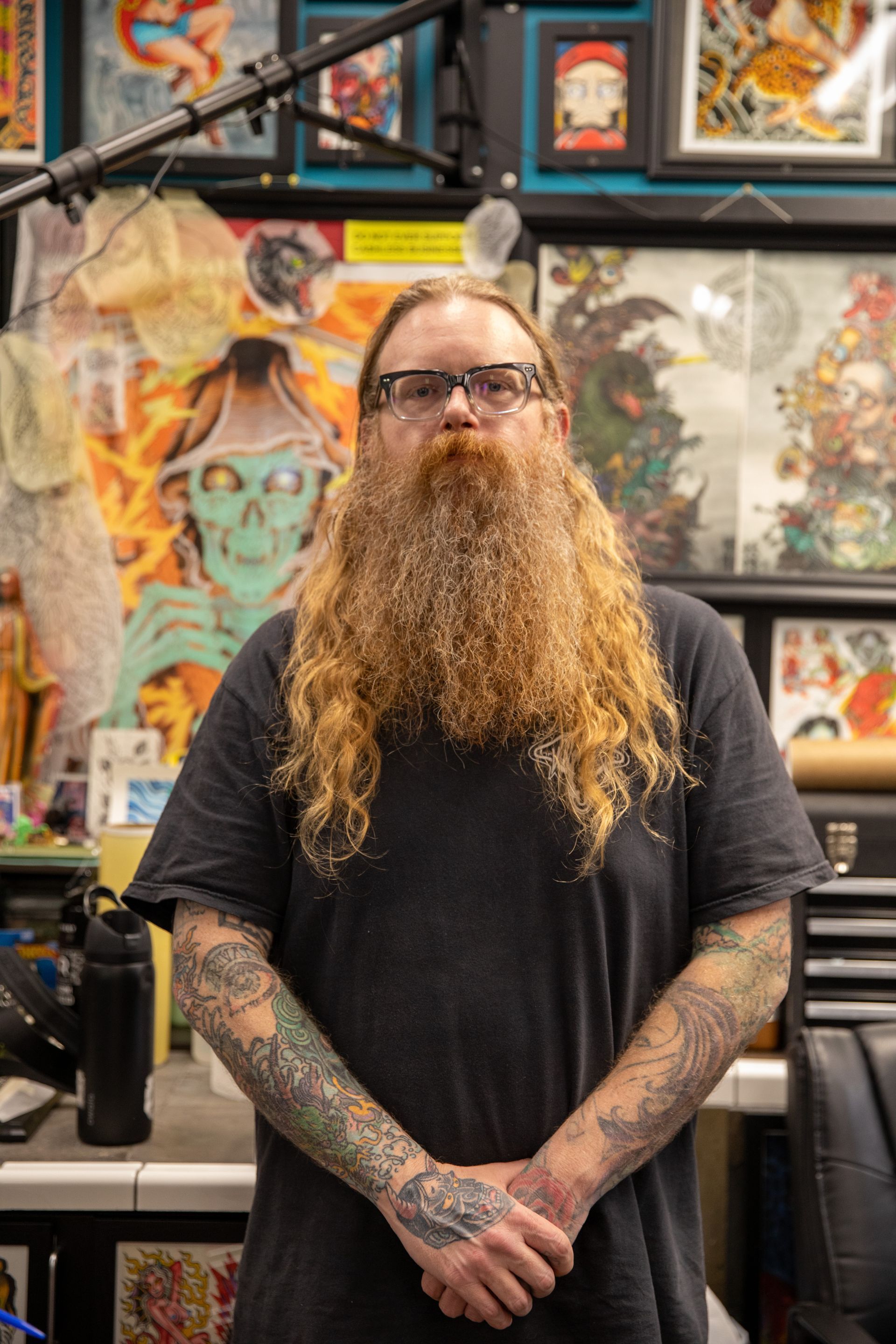 Person with long red beard and tattooed arms standing in a tattoo studio, framed art behind them