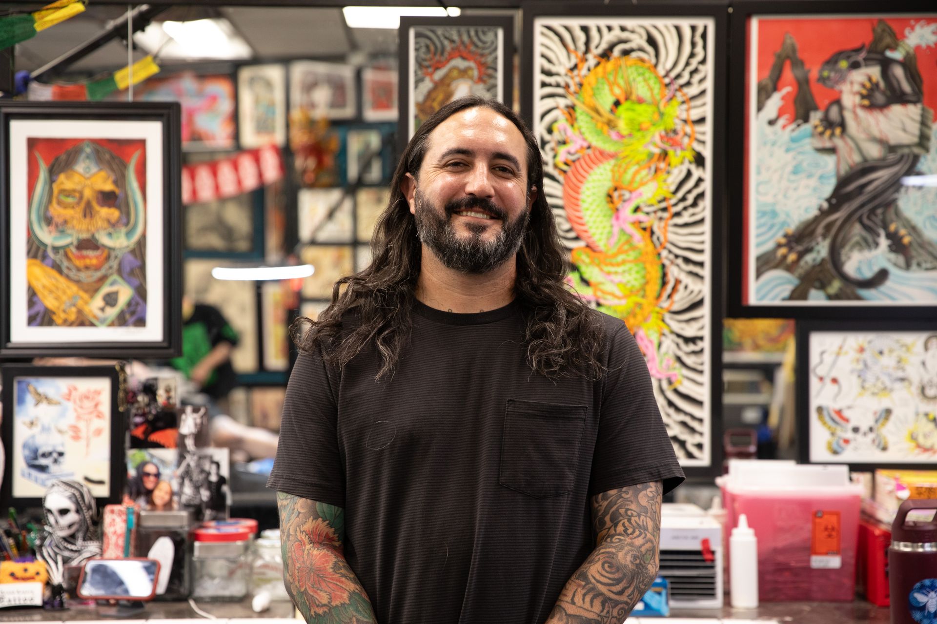 Smiling tattoo artist in a shop, standing before colorful framed artwork and tattoo designs.