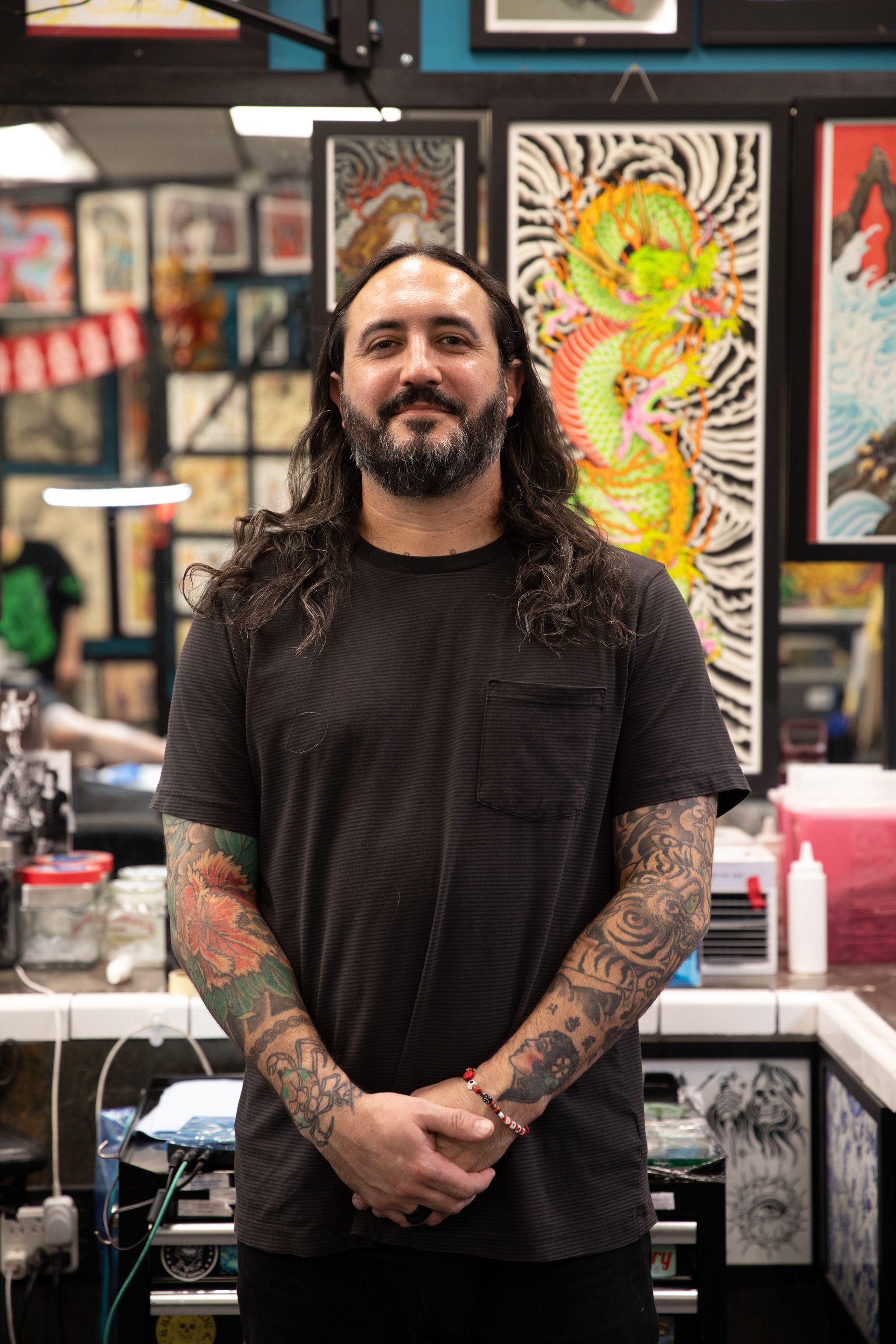 Tattooed person in a black shirt standing in a colorful tattoo studio with framed artwork behind them