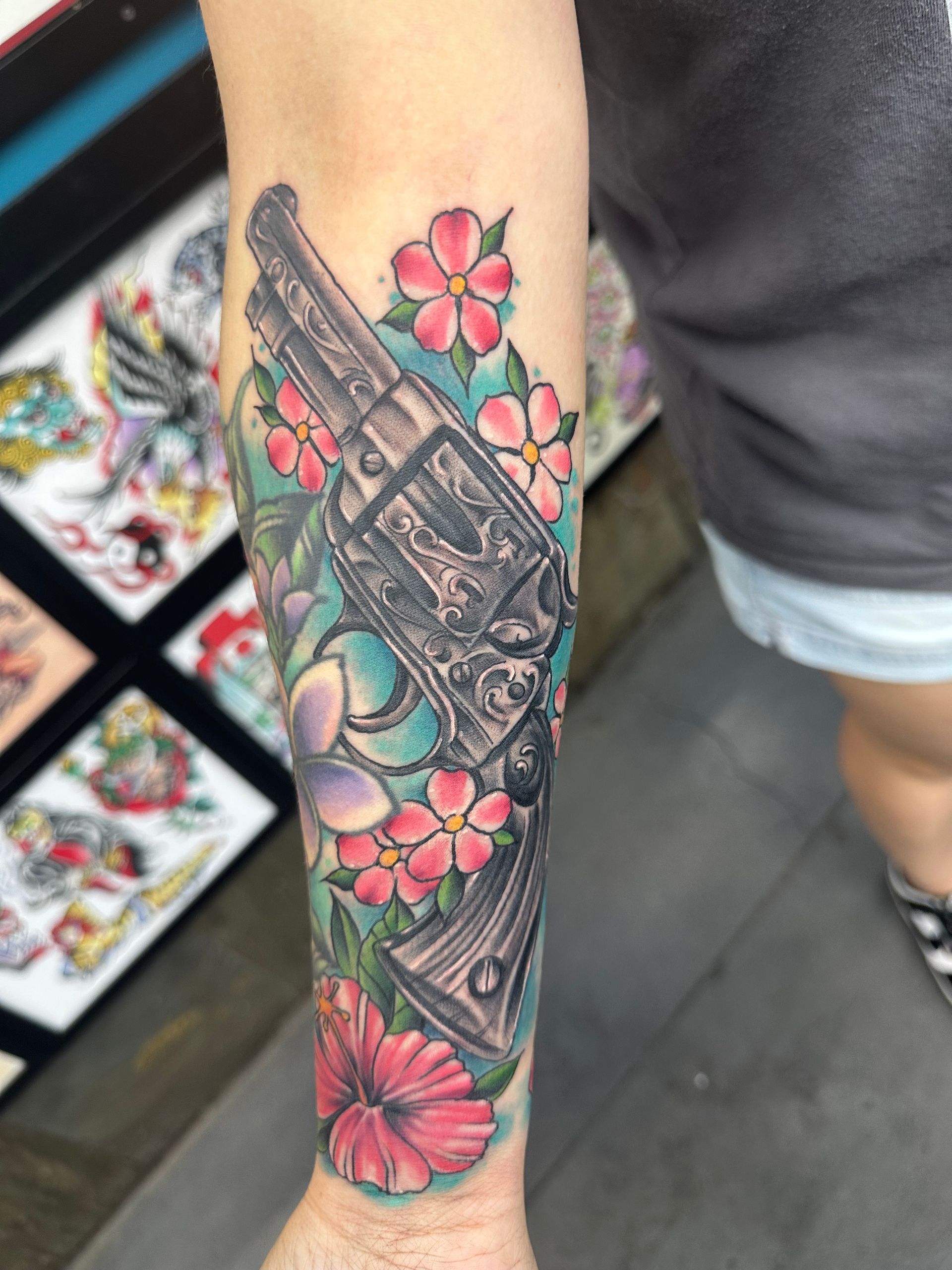 Forearm tattoo of a revolver surrounded by pink and green flowers