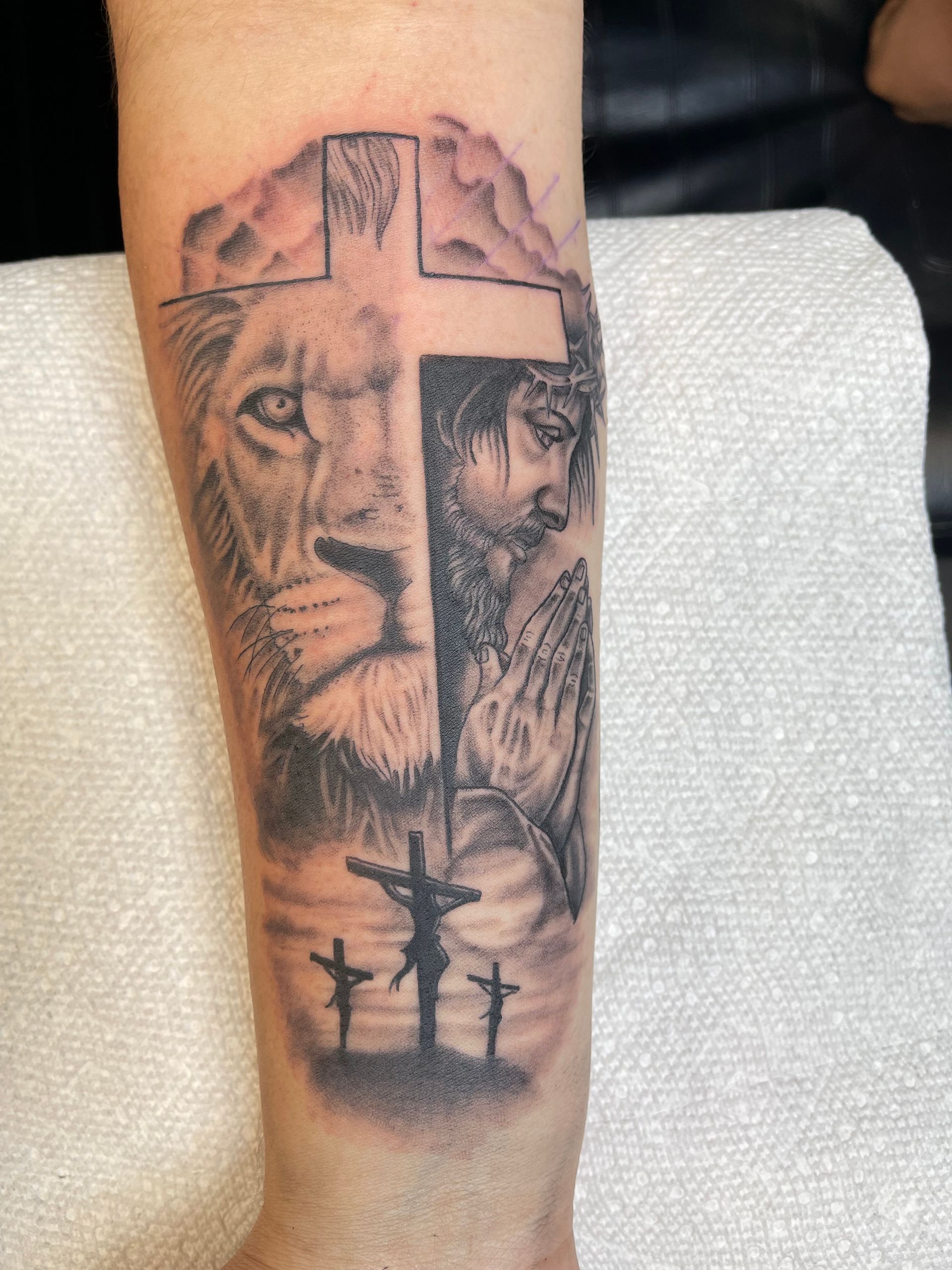Black-and-gray forearm tattoo of a lion’s face with a cross and silhouetted figures below