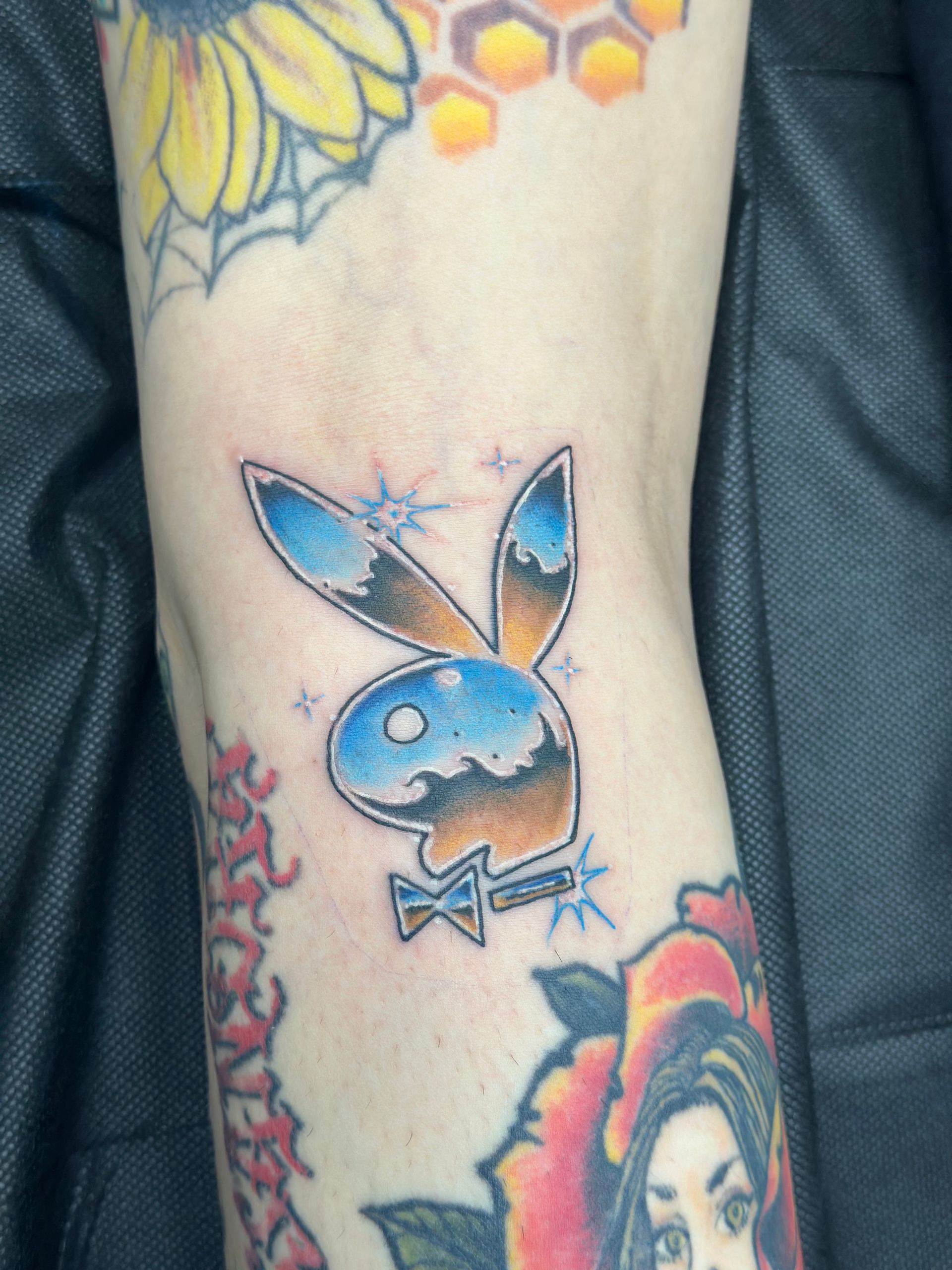 Blue cartoon bunny tattoo with pink sparkles on a person's forearm, surrounded by colorful floral tattoos