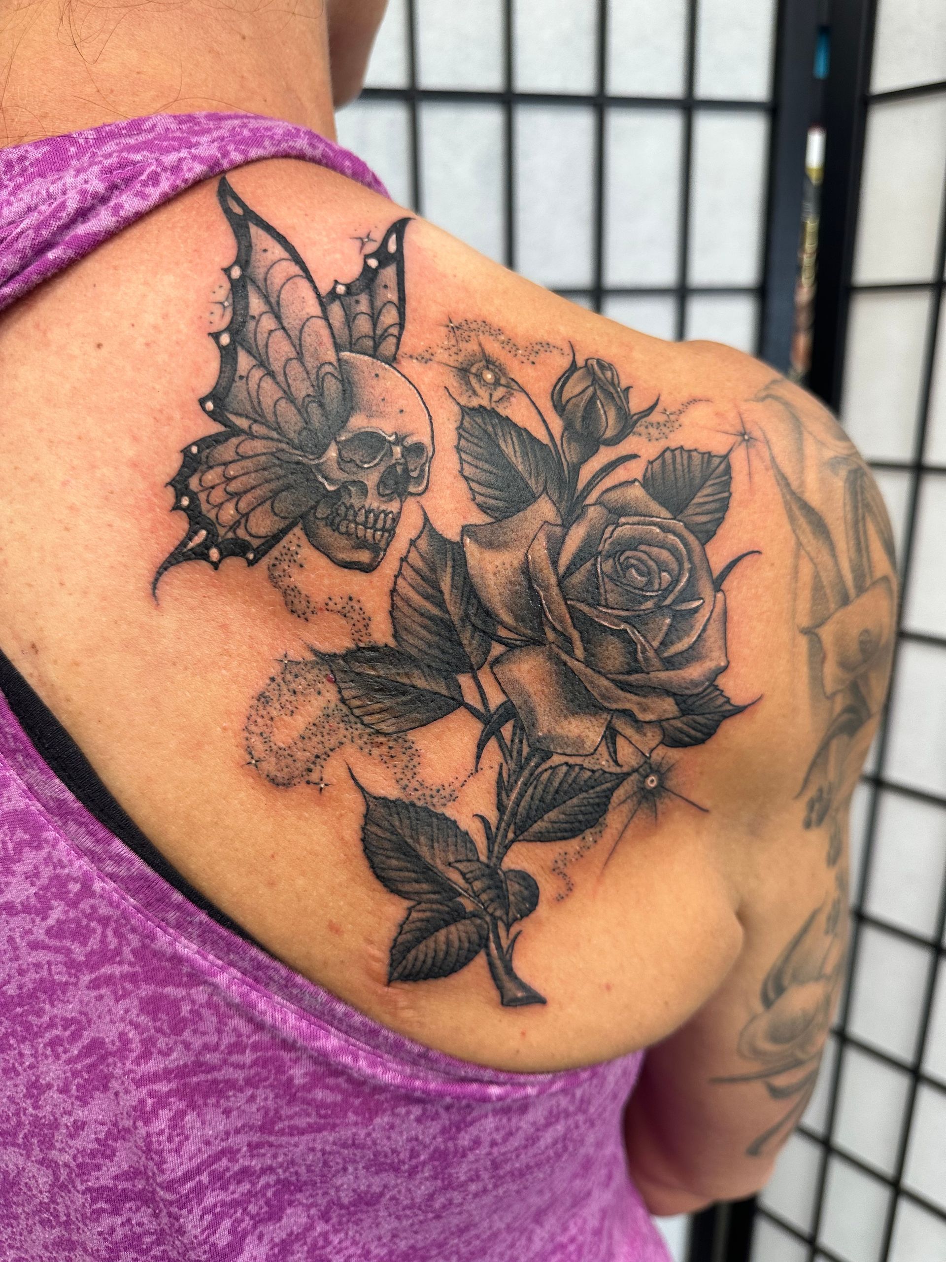 Shoulder tattoo of roses, butterflies, and leaves in black and gray on a person in a purple top